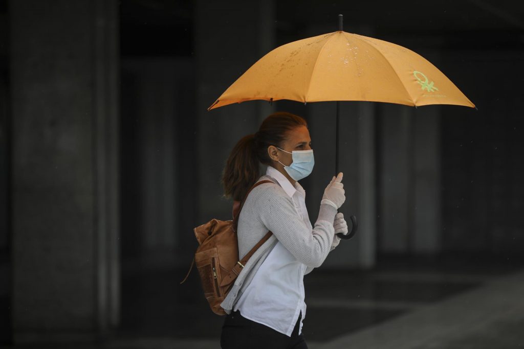 Una mujer protegida con mascarilla y guantes se resguarda de la lluvia bajo su paraguas Moncloa