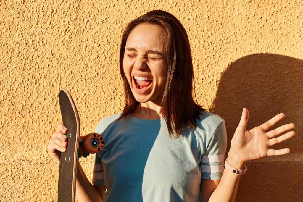 Calor y violencia: ¿Cómo los episodios de calor afectan a nuestro comportamiento y desatan conflictos? 5 Moncloa foto de mujer de pelo oscura emocionada con camiseta azul de pie contra la pared amarilla al aire libre y gritando alegremente sosteniendo longboard en las manos expresando felicidad Moncloa